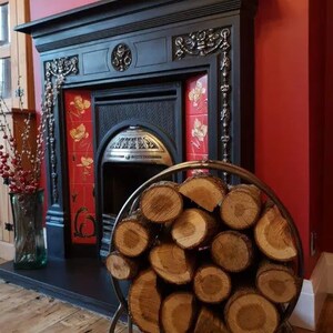 Puede incluir: Una chimenea de metal negro con un marco de azulejos rojos y blancos. Un porta-le&ntilde;a redondo de metal con troncos apilados se encuentra frente a la chimenea.