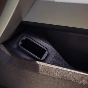 May include: A black rectangular object, possibly a phone or small device, is nestled within a dark compartment. The compartment is part of a larger, textured, dark gray structure, possibly a car interior. The overall scene is dimly lit.