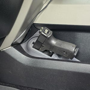 May include: A black handgun is secured in a gray, custom-fit holder within a vehicle's interior. The holder is integrated into the dashboard, providing a discreet storage solution. The image showcases the firearm's details and the holder's design.