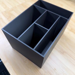 May include: A dark gray rectangular organizer with multiple compartments. The organizer is made of a matte material and is designed to hold various items. The compartments are divided by vertical dividers, creating separate sections for storage. The organizer sits on a light-colored wooden surface.