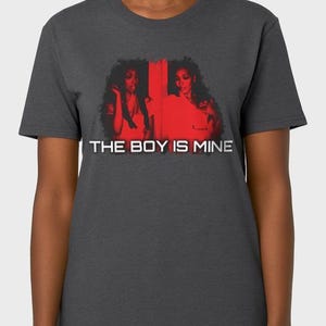 Pode incluir: T-shirt cinzenta escura com um gráfico vermelho de duas mulheres e o texto branco "THE BOY IS MINE". A camisa é feita de um material macio e tem gola redonda.