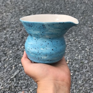 May include: A small, light blue ceramic jug with a speckled glaze. The jug has a rounded body and a slightly flared lip.