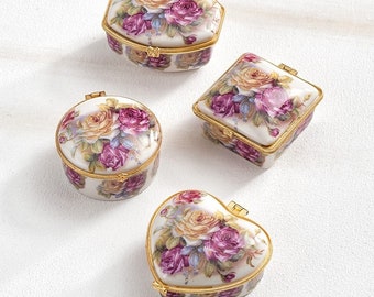 May include: Four porcelain trinket boxes with floral designs. The boxes are in the shapes of a heart, a square, a circle, and a hexagon. Each box has a gold trim and a hinged lid.