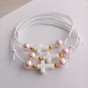 Könnte beinhalten: Drei weiße Armbänder mit Kreuzanhängern, rosa und goldenen Perlen. Die Armbänder sind aus weißem Band gefertigt und miteinander verbunden.