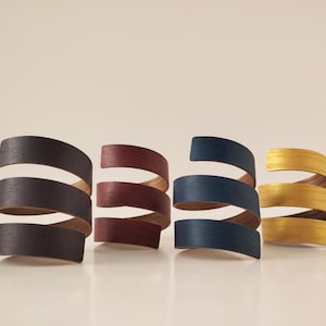 Könnte beinhalten: Vier Holzringe, jeder in einer anderen Farbe lackiert: dunkelblau, burgunderrot, gold und schwarz. Die Ringe sind übereinander gestapelt und erzeugen so einen Spiraleffekt.