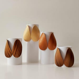 May include: Four pairs of wooden teardrop earrings in different wood tones. The earrings are displayed on white stands.