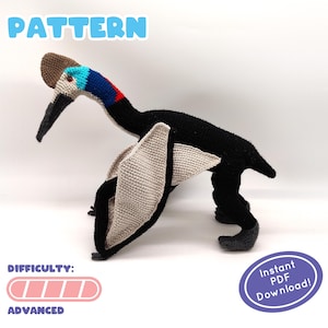 Puede incluir: Patrón de crochet para un pterodáctilo negro y gris con una cresta azul y roja. El patrón está etiquetado como "Avanzado" e incluye un botón "Descarga instantánea de PDF".