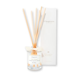 May include: A clear glass reed diffuser with wooden reeds and a white ribbon. The label reads "Chapters White Flowers". A white box with the "Chapters" logo and text is behind the diffuser. The product is designed for home fragrance.