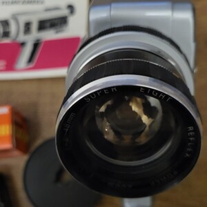 以下が含まれることがあります： 大きなレンズが付いたシルバーのSuper Eight Reflexフィルムカメラ。レンズは黒とシルバーで、「SUPER EIGHT」と「REFLEX」の文字が印刷されています。レンズの測定値は48mmです。「razzoom」という文字が入った箱が背景にあります。