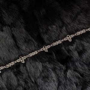 May include: A silver chain necklace with a delicate design and small decorative elements. The necklace is displayed on a dark, textured surface, creating a contrast that highlights its intricate details.