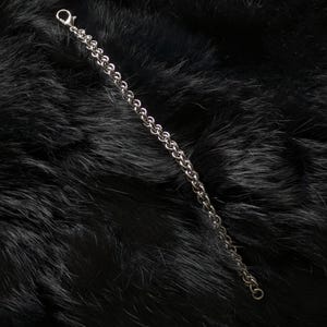 May include: A silver-colored chain bracelet with a lobster clasp. The bracelet is made of small, interconnected links. The background is a dark, textured surface.
