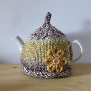 May include: A white teapot with a knitted tea cosy. The cosy is in shades of yellow, purple, and white, with a large yellow flower and a wooden button. The cosy covers the teapot and handle.