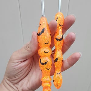 May include: Two orange crochet hooks with silver metal hooks. The handles are decorated with black bat silhouettes and glitter. The hooks are held in a hand, and are designed for crafting.