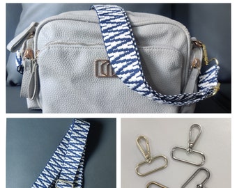 May include: A white and blue striped fabric bag strap with gold metal hardware. The strap is shown with two silver swivel clasps.