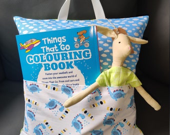 Pode incluir: Uma almofada azul e branca com um bolso que contém um livro intitulado "Things That Go Coloring Book" e um animal de pelúcia. A almofada tem uma alça branca e uma estampa de dinossauro.