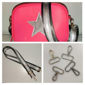 May include: A pink crossbody bag with a silver star and black and silver striped strap. The bag comes with two silver and gold metal clasps.