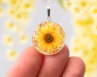 May include: A silver chain necklace with a round pendant featuring a preserved sunflower encased in clear resin. The sunflower has yellow petals and a brown center.