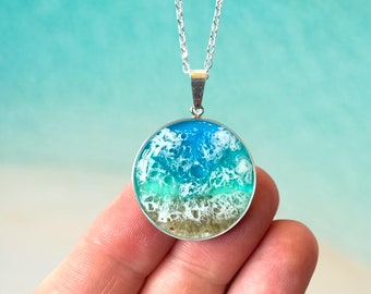 Può includere: Una collana con catena d'argento con un ciondolo rotondo che presenta un design di onda oceanica blu e verde con schiuma bianca e sabbia sul fondo.