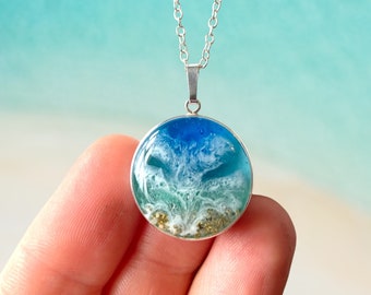 May include: A silver chain necklace with a round pendant featuring a blue, green, and white resin ocean wave design with gold glitter accents.