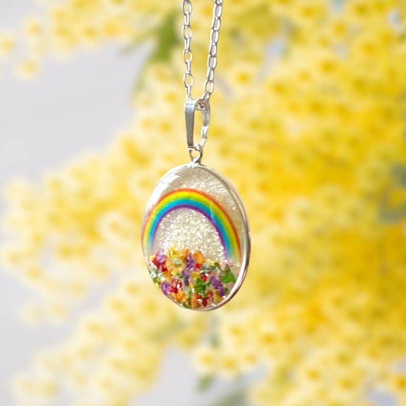 May include: A silver chain necklace with a clear oval pendant featuring a rainbow and colorful flowers pressed inside. The pendant is filled with glitter.