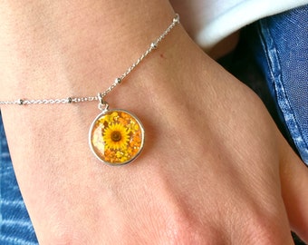 May include: A silver bracelet featuring a round pendant with a pressed sunflower design. The pendant is filled with yellow and orange flower petals. The bracelet is on a wrist.