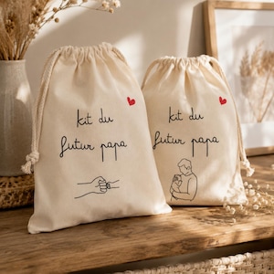 Peut inclure: Deux sacs en coton écru avec cordon de serrage, portant l'inscription noire "kit du futur papa" et un petit cœur rouge. L'un présente un dessin au trait d'un check de poings, l'autre un dessin d'une personne tenant un bébé.