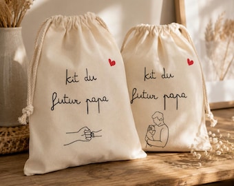Puede incluir: Dos bolsas de algodón color crema con cordón, con el texto negro "kit du futur papa" y un pequeño corazón rojo. Una bolsa muestra un dibujo lineal de un choque de puños, la otra un dibujo de una persona sosteniendo a un bebé.