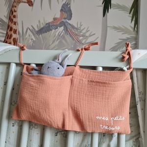 Può includere: Un organizzatore in tessuto color terracotta con tre tasche è appeso a una culla. L'organizzatore ha il testo "mes petits trésors" ricamato sulla tasca inferiore. Un coniglietto di peluche grigio è nella tasca superiore.