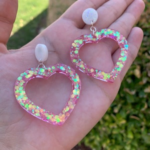 May include: A pair of pink heart-shaped earrings with iridescent glitter embedded in the resin. The earrings have a white pearl-like accent at the top.