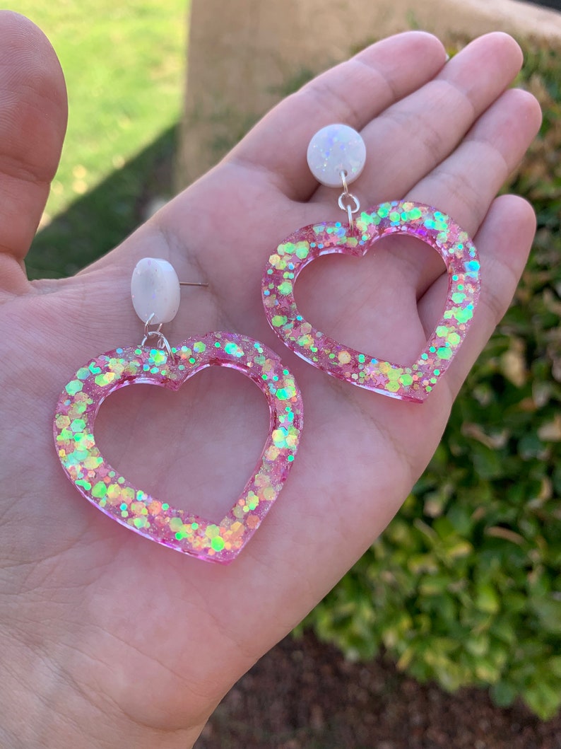 May include: A pair of pink heart-shaped earrings with iridescent glitter embedded in the resin. The earrings have a white pearl-like accent at the top.