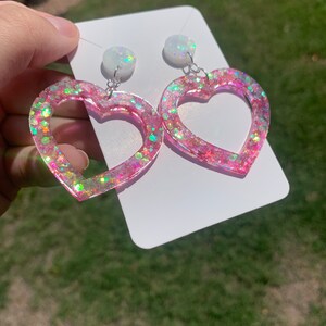 May include: A pair of pink heart-shaped earrings with iridescent glitter. The earrings have a white opal-like charm at the top.