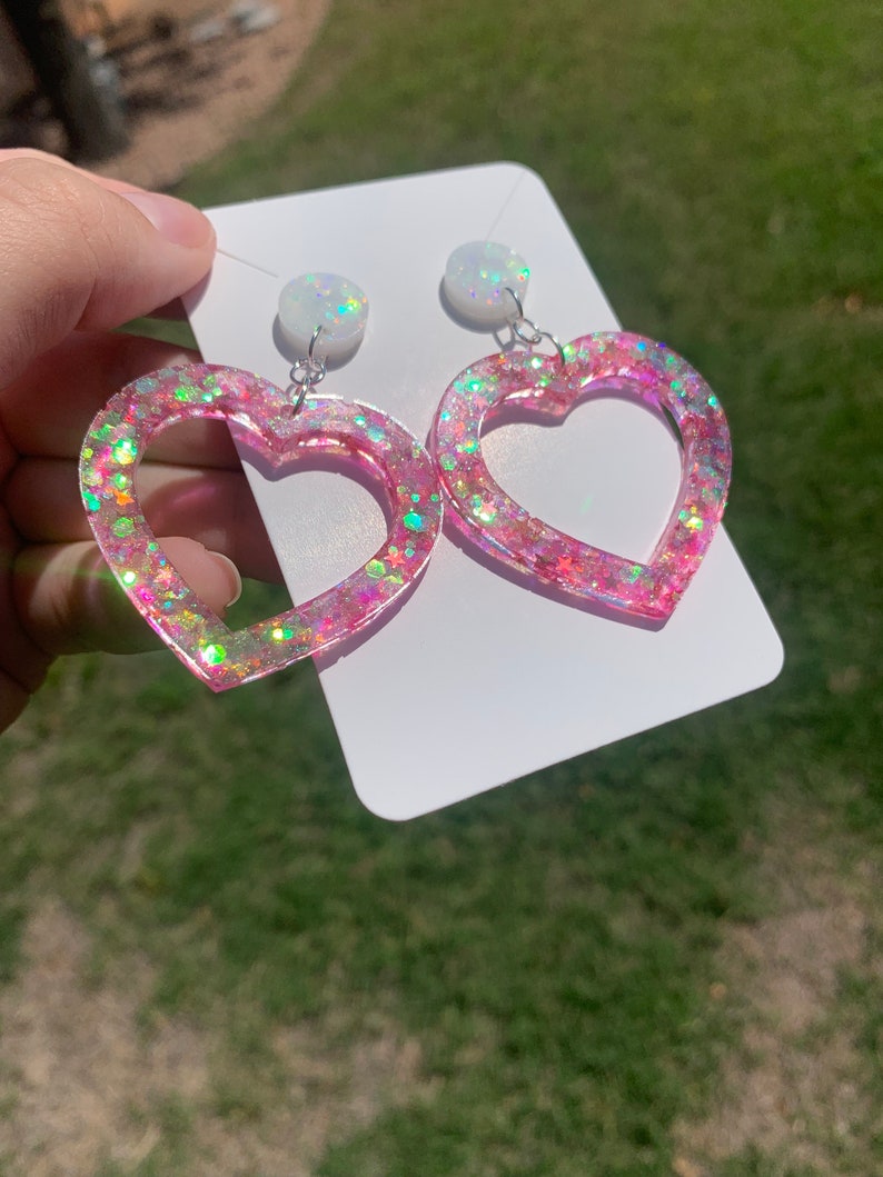 May include: A pair of pink heart-shaped earrings with iridescent glitter. The earrings have a white opal-like charm at the top.