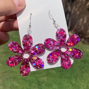 May include: A pair of pink glitter flower earrings with silver hooks. The flowers have a six-petal design and are filled with iridescent glitter.