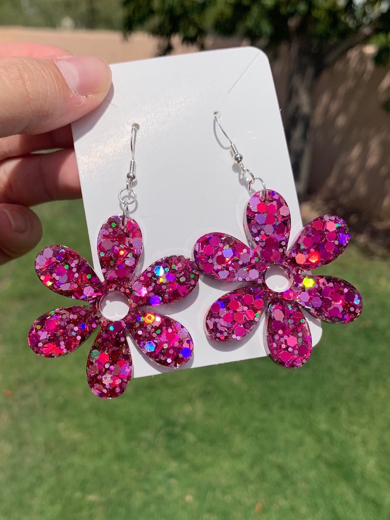May include: A pair of pink glitter flower earrings with silver hooks. The flowers have a six-petal design and are filled with iridescent glitter.