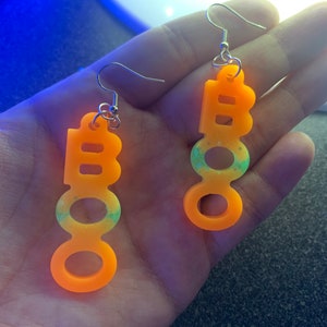 May include: A pair of orange acrylic earrings spelling out "BOO" in a playful font. The earrings have silver hooks and a gradient of green and blue glitter. The earrings are designed for Halloween.