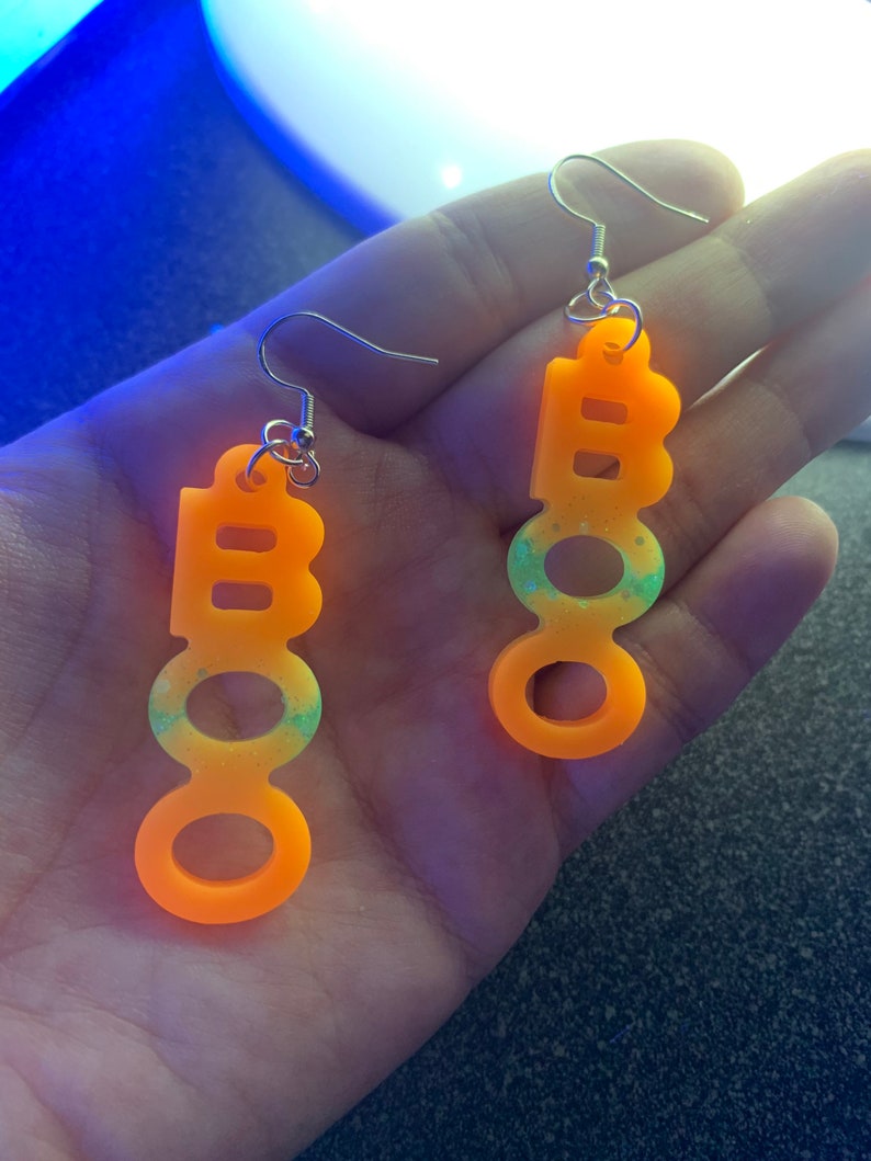 May include: A pair of orange acrylic earrings spelling out "BOO" in a playful font. The earrings have silver hooks and a gradient of green and blue glitter. The earrings are designed for Halloween.