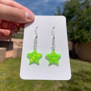 May include: A pair of star-shaped earrings. Each earring features a bright neon green color with glitter accents. The earrings hang from silver-colored chains and hooks, presented on a white card.