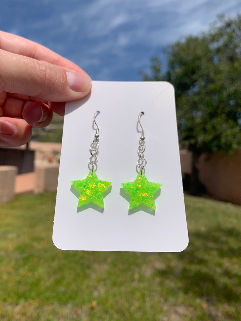 May include: A pair of star-shaped earrings. Each earring features a bright neon green color with glitter accents. The earrings hang from silver-colored chains and hooks, presented on a white card.