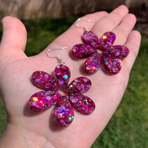 May include: A pair of pink flower-shaped earrings with iridescent glitter embedded in the resin. The earrings have silver hooks.