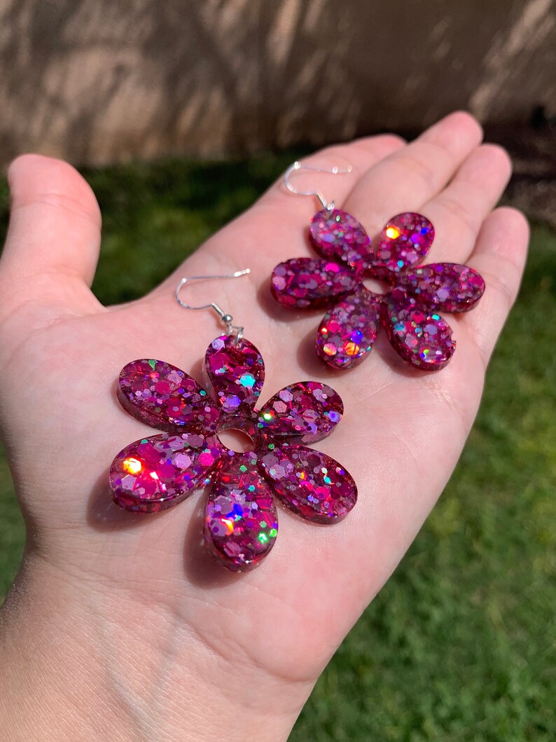 May include: A pair of pink flower-shaped earrings with iridescent glitter embedded in the resin. The earrings have silver hooks.