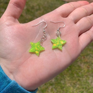 May include: A pair of star-shaped earrings. Each earring features a bright green, glitter-infused star pendant with silver-colored ear wires. The earrings are held in a hand against a blurred green background.