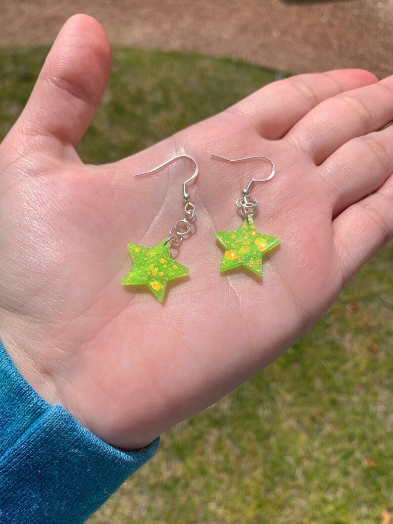 May include: A pair of star-shaped earrings. Each earring features a bright green, glitter-infused star pendant with silver-colored ear wires. The earrings are held in a hand against a blurred green background.