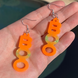 May include: A pair of orange "BOO" earrings with silver hooks. The earrings have a gradient effect, transitioning from orange to a light green with glitter. The earrings are designed for Halloween.