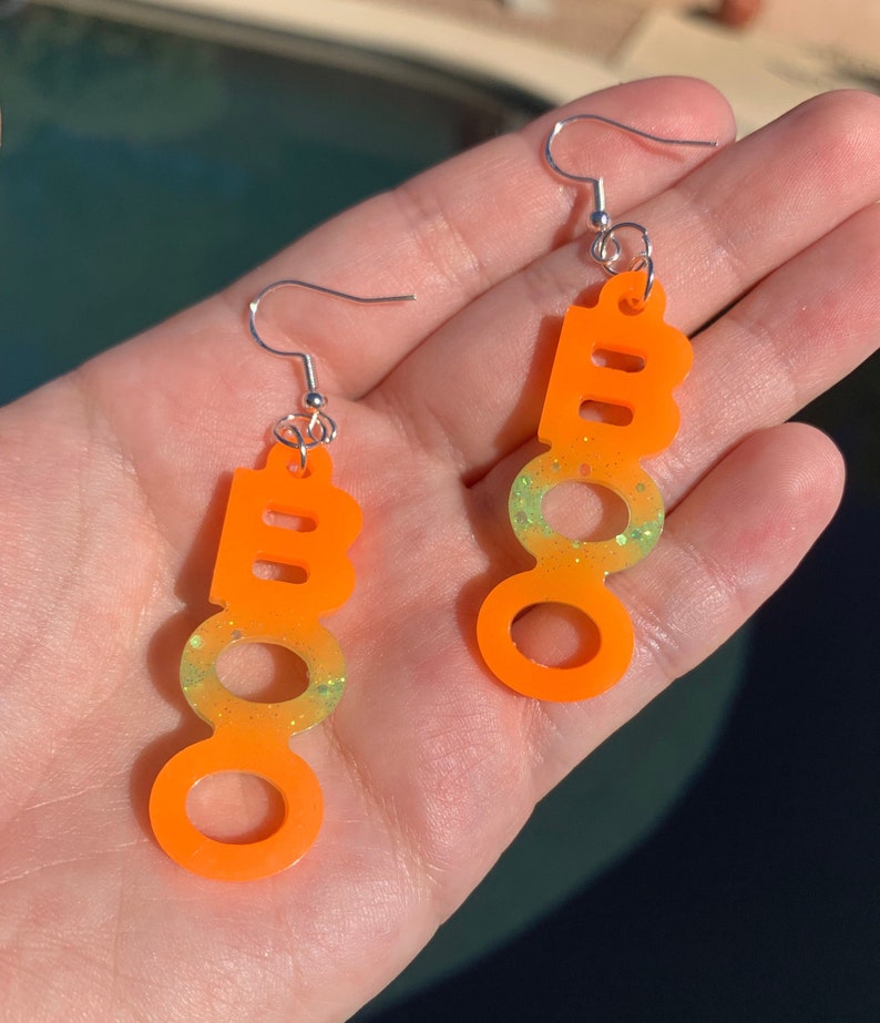 May include: A pair of orange "BOO" earrings with silver hooks. The earrings have a gradient effect, transitioning from orange to a light green with glitter. The earrings are designed for Halloween.