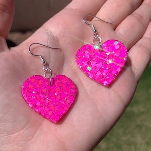 May include: A pair of vibrant pink heart-shaped earrings with silver hooks. The earrings are embellished with glitter, reflecting light. These earrings are a cheerful and playful accessory.