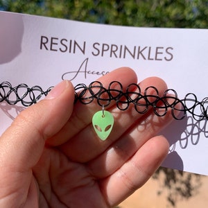May include: A black choker necklace with a green alien charm. The charm is made of resin and has a glittery finish. The choker is made of elastic and has a stretchy fit.