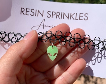 May include: A black choker necklace with a green alien charm. The charm is made of resin and has a glittery finish. The choker is made of elastic and has a stretchy fit.