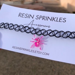 May include: A black choker necklace with a pink glitter spider charm. The charm has green glitter accents for the eyes. The necklace is on a white card with the text "RESIN SPRINKLES Accessories RESINSPRINKLES.ETSY.COM".