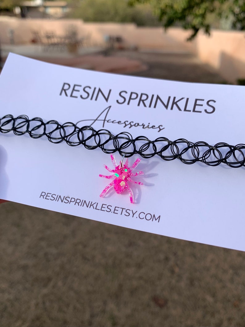 May include: A black choker necklace with a pink glitter spider charm. The charm has green glitter accents for the eyes. The necklace is on a white card with the text "RESIN SPRINKLES Accessories RESINSPRINKLES.ETSY.COM".