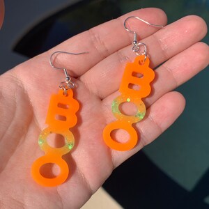 May include: A pair of orange "BOO" earrings with silver hooks. The earrings have a gradient effect, transitioning from orange to yellow-green with glitter accents. The earrings are designed for Halloween.